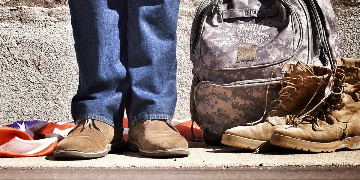 Veteran standing with a camouflage backpack, boots, and an American flag close to them